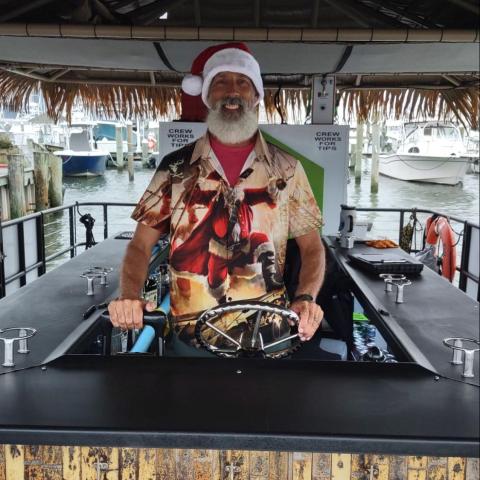 Man in Santa hat and festive shirt stands at boat helm under a thatched roof.