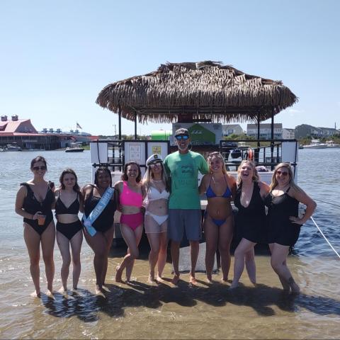 Group of people in swimwear standing in the water in front of a tiki bar on a sunny day.