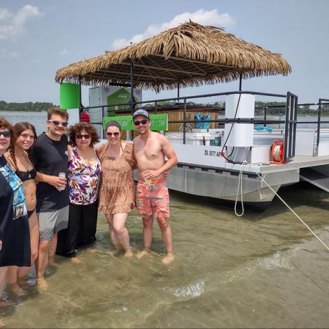 Group of people standing in shallow water by a tiki hut boat on a sunny day.