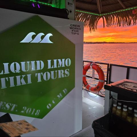 Tiki boat bar interior with sunset view over water, green sign, and life ring.