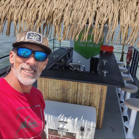 Man in red shirt and cap on a tiki-themed boat with bar seating.