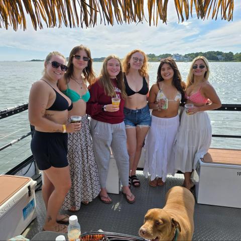 Six women in summer attire on a boat with drinks, a dog, and water view.