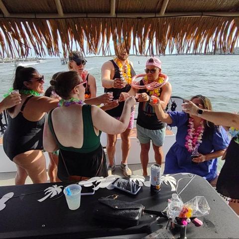 Group of people on a boat toasting drinks, wearing leis and summer attire.