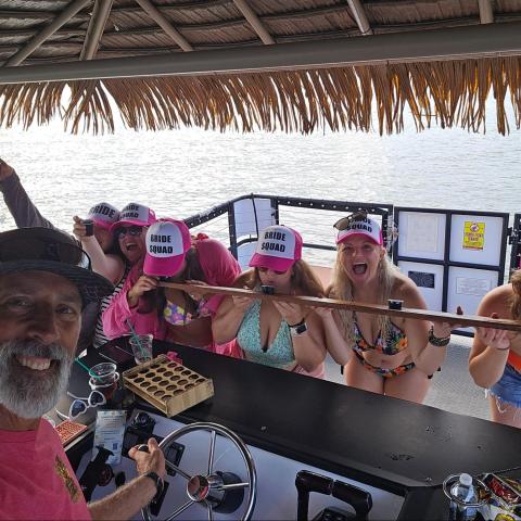 Group on a boat, women in 'Bride Squad' hats doing a shot ski, man at the helm in a pink shirt.