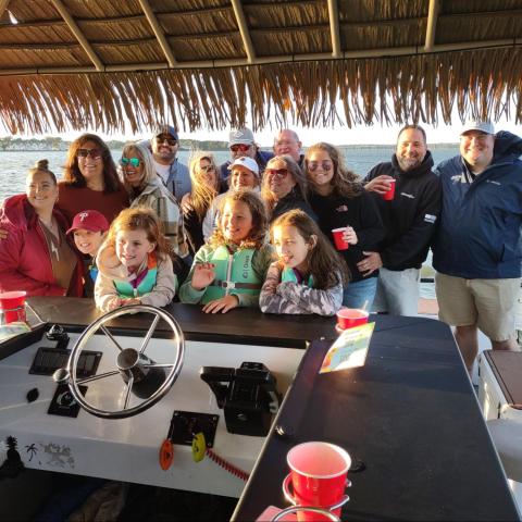 Group of people smiling on a boat under a thatched roof, holding red cups, with water in the background.