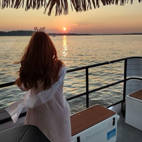 Woman in bride sash stands on boat at sunset, overlooking water.