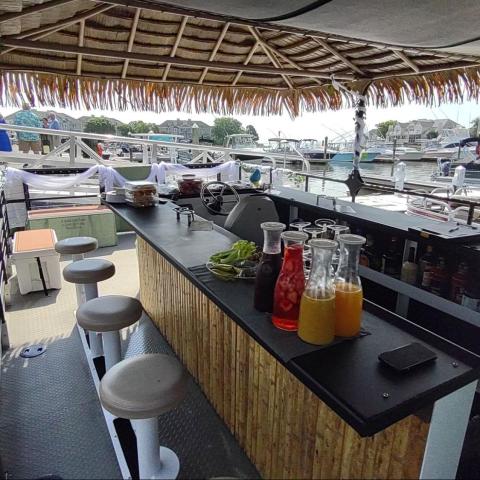 Outdoor bar on a floating platform with drinks and stools under a thatched roof by a marina.