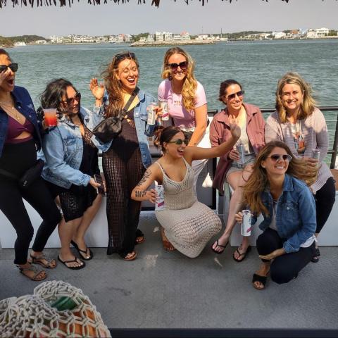 Group of nine women posing on a boat with drinks, ocean and houses in the background.