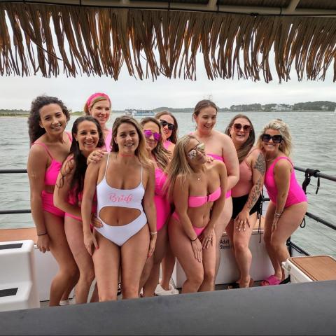 Group of women in pink swimsuits on a boat, one wearing a white 'Bride' swimsuit.