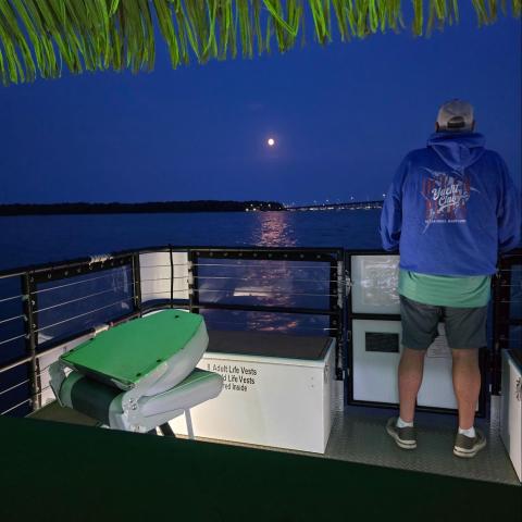 Person in hoodie on boat at night, gazing at full moon over the water.