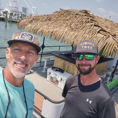 Two men on a dock next to a boat with a thatched roof on a sunny day.