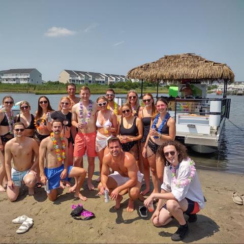 Group of people posing on a sandy beach near a tiki-style boat, wearing swimwear and colorful leis.