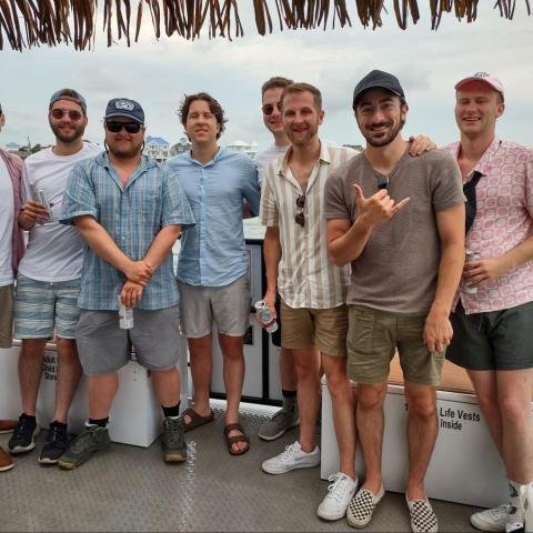 Eight men posing on a boat deck with water and buildings in the background.