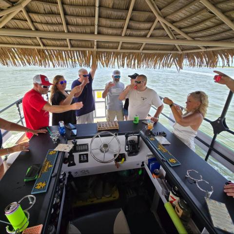 People having drinks on a tiki bar boat with a thatched roof, surrounded by water.