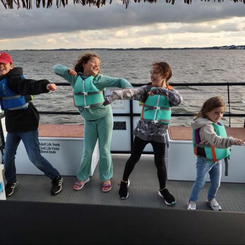Four kids in life jackets dancing on a boat with a cloudy sky and water in the background.