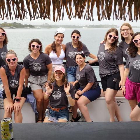 Group of women in matching shirts and heart-shaped sunglasses on a boat, smiling under thatched roof.