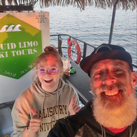 Two people smiling on a boat with a tiki roof, ocean in background, and tour sign visible.