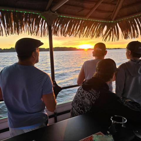 Four people on a boat watching a sunset over the water under a thatched roof.