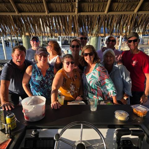 Group of smiling people posing under a thatched roof by the marina at sunset.