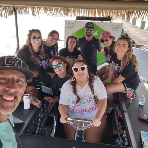 Group of people smiling on a boat with drinks, near a dock.