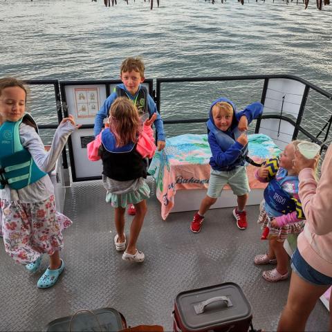 Children wearing life jackets dancing on a boat with a woman holding a drink, over water.