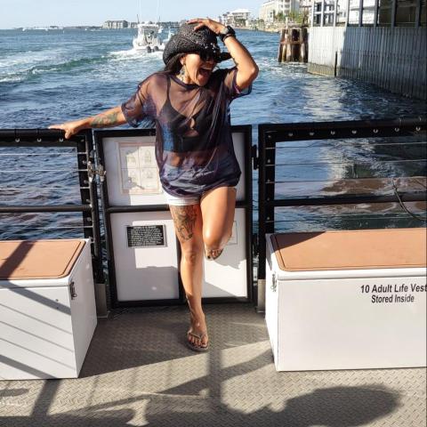 Person posing on a boat under a bridge, with ocean and buildings in the background.