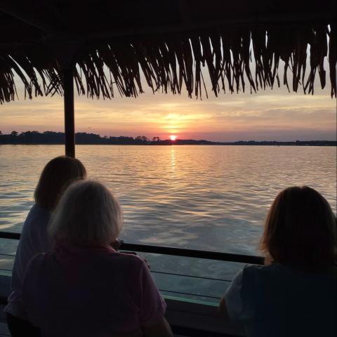 Three people watching a sunset over a lake, framed by a thatched roof.