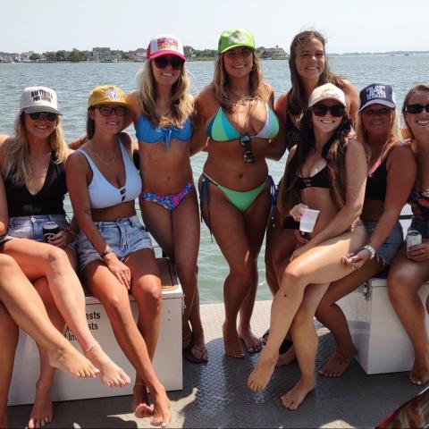 Group of women in swimwear and hats sitting on a boat, with water and distant coastline in the background.