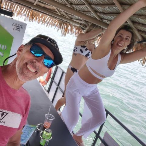 Group on a tiki boat, man in sunglasses, woman posing, ocean in background.