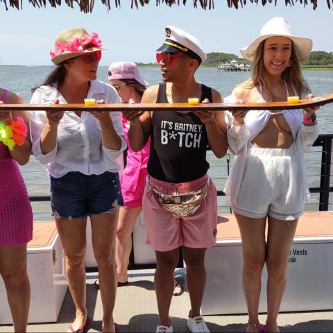 Group of people on a boat holding a plank with shot glasses, under a tiki roof, with water in the background.