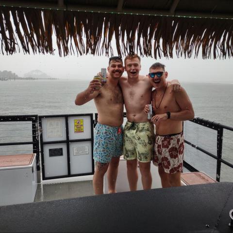 Three men in swimwear posing happily on a boat under a thatched roof, with a rainy sea view.